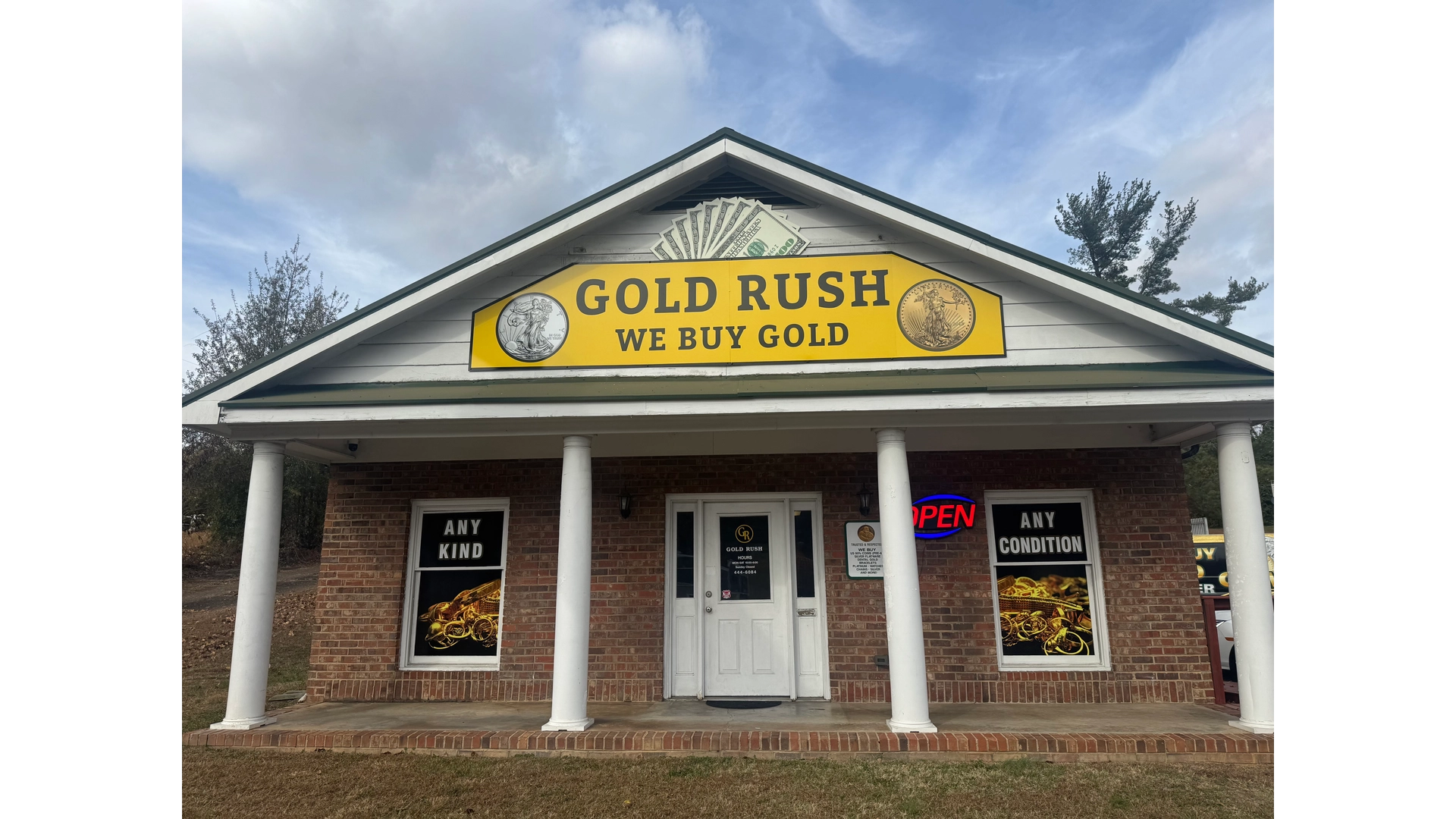 Brick storefront with white pillars featuring a yellow sign that reads 'GOLD RUSH WE BUY GOLD' and an open neon sign on the door.