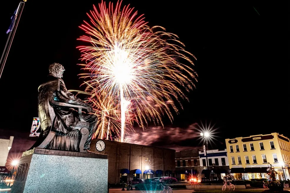 Fireworks lighting up the night sky behind a statue seated on a pedestal in a town square.