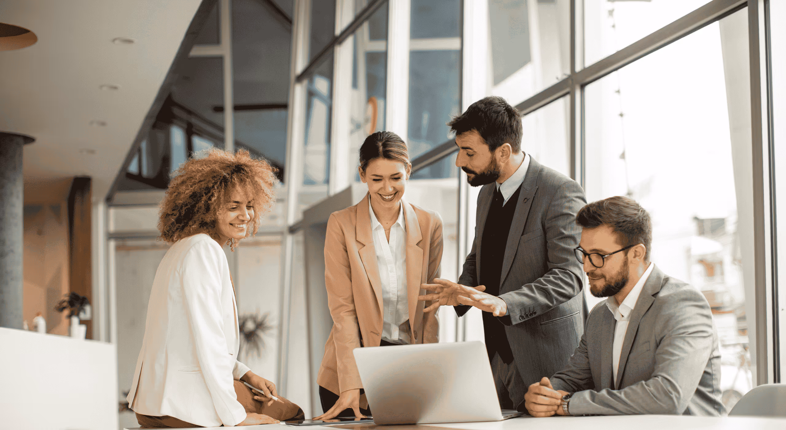 Four business professionals collaborating around a laptop in a modern office with large windows.