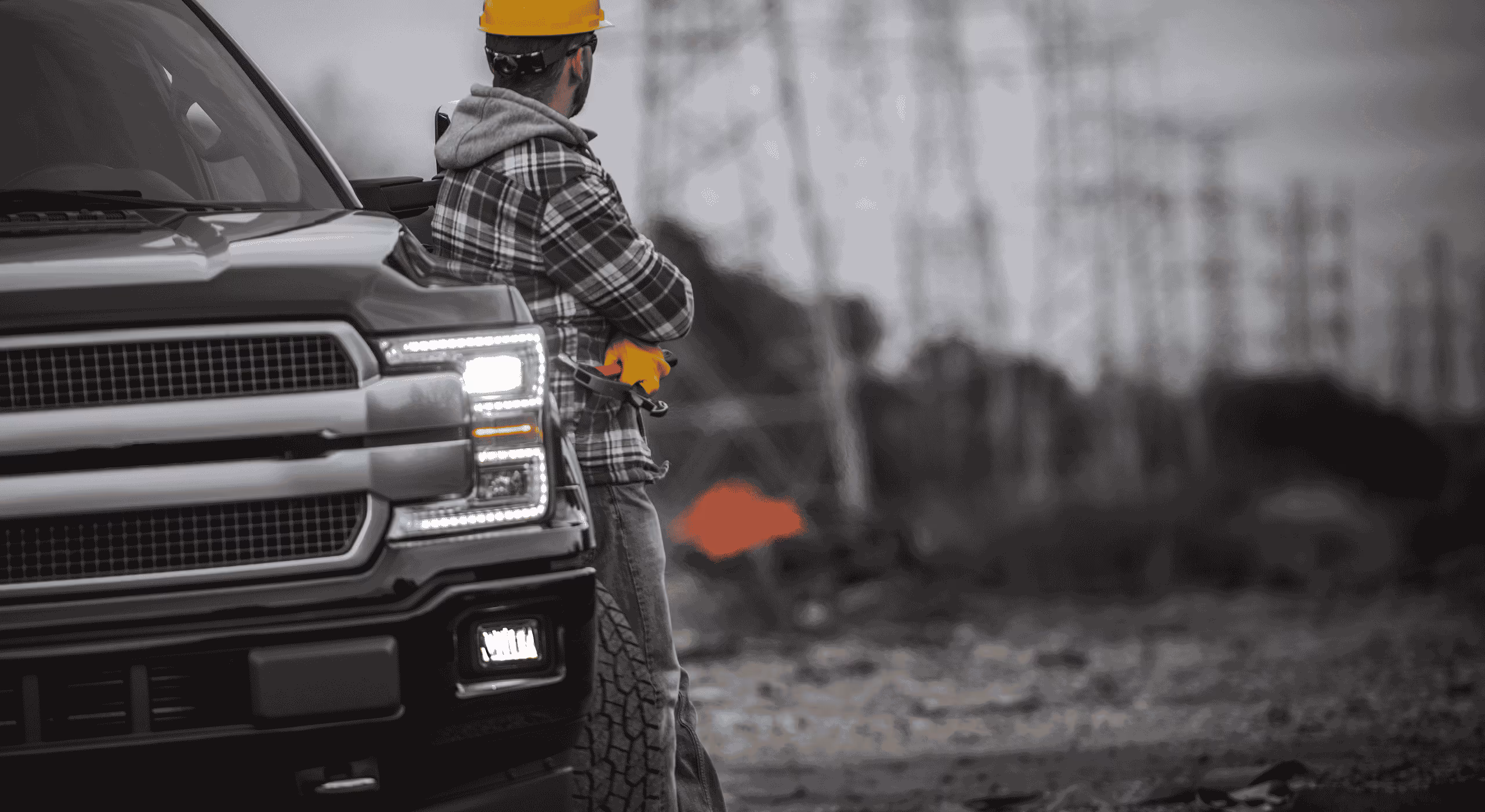 Construction worker wearing a hard hat and gloves leaning against a black pickup truck in a rugged outdoor setting.
