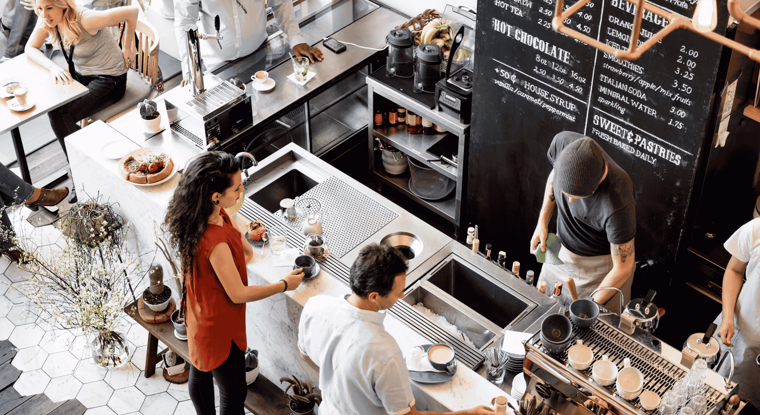 Busy cafe counter with barista preparing drinks and customers ordering and sitting at tables.