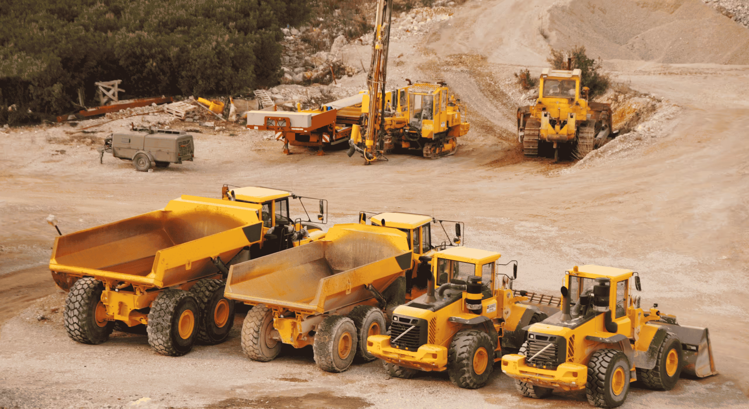 Row of large yellow construction vehicles including dump trucks and loaders in a quarry with dirt and rocky terrain.