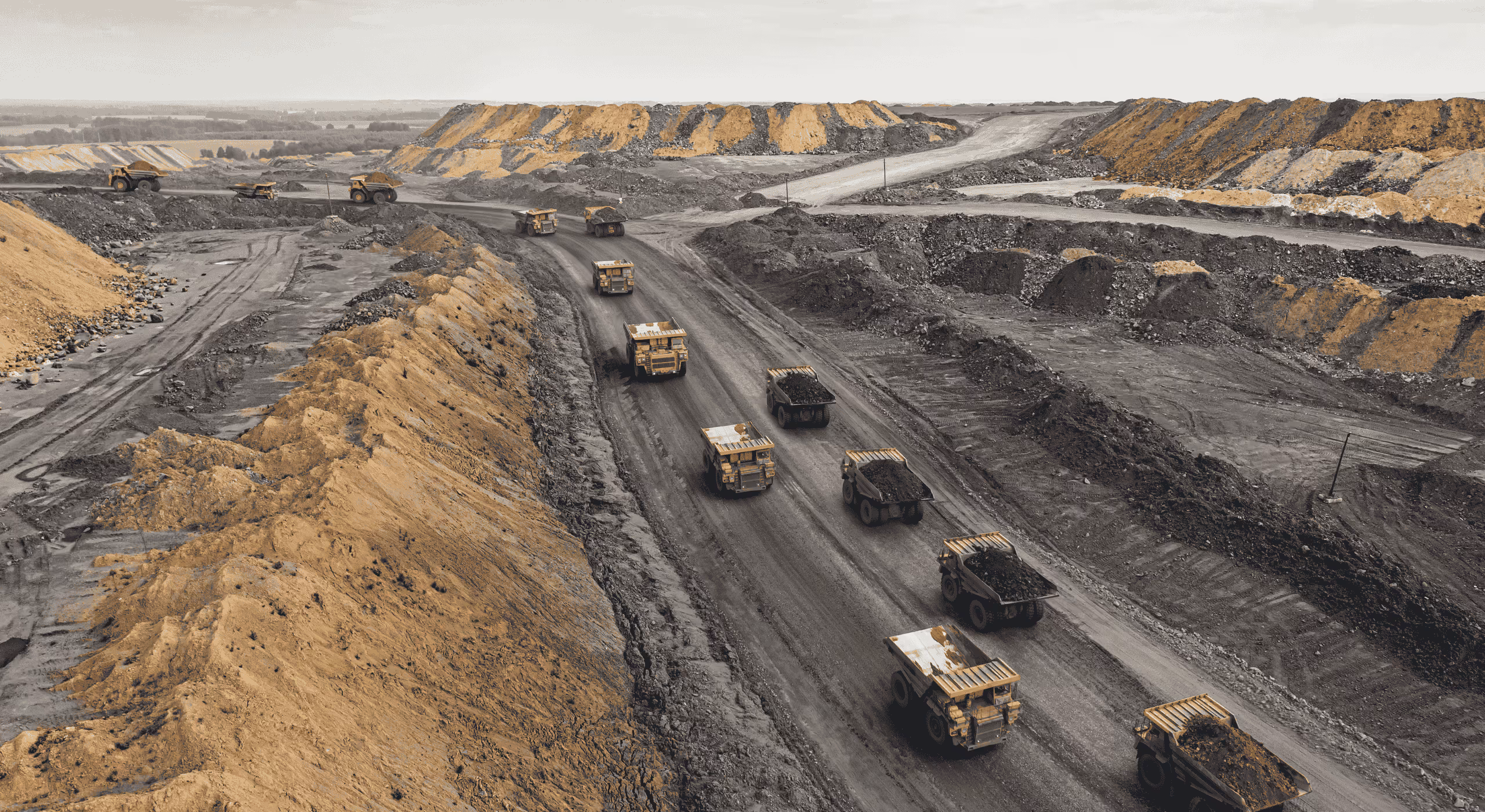 Aerial view of multiple large mining trucks transporting soil and minerals on a winding dirt road in an open-pit mine.