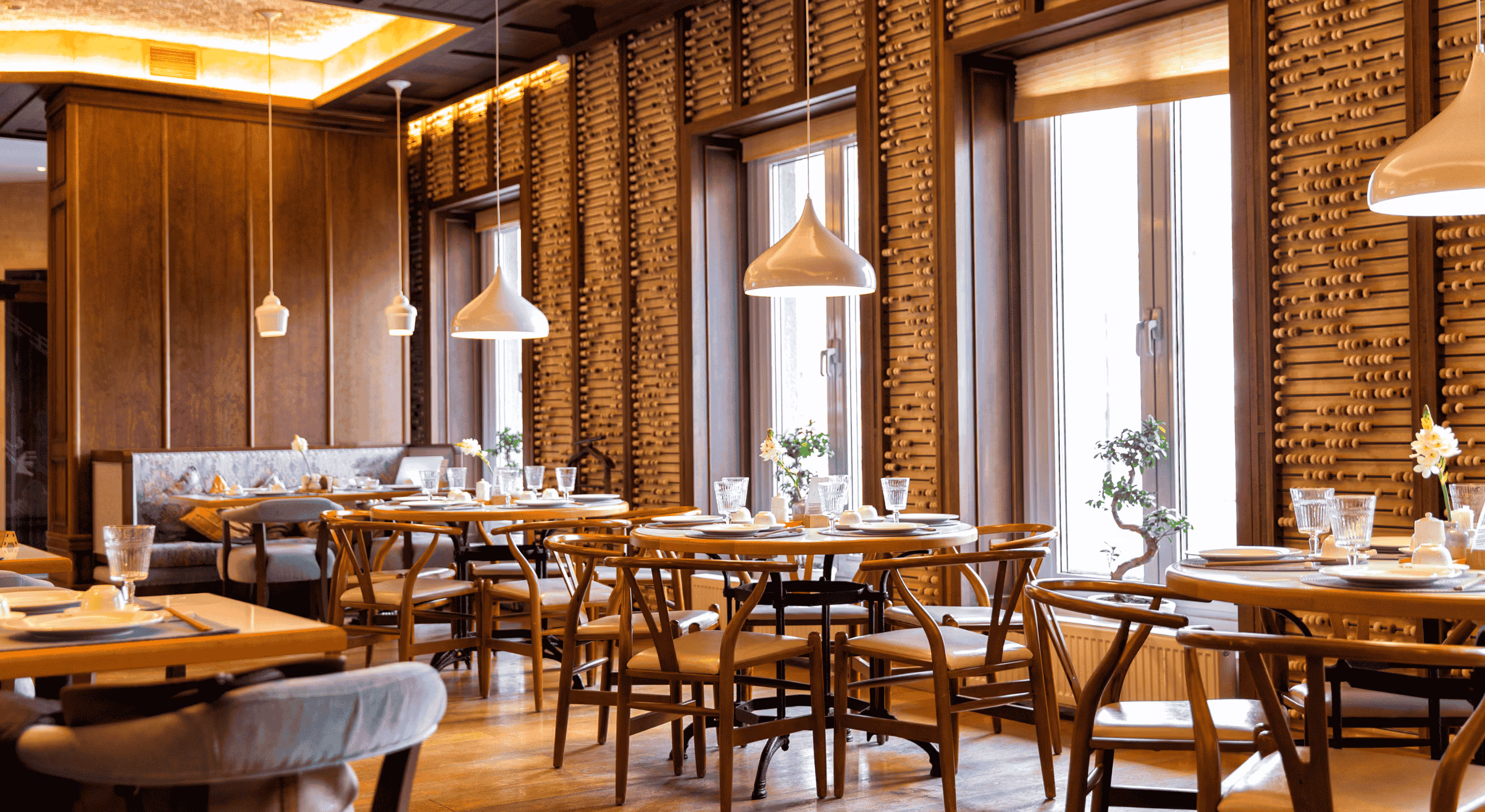 Elegant restaurant interior with wooden chairs and round tables set with glassware and plates, pendant lights, and large windows with decorative wooden wall panels.
