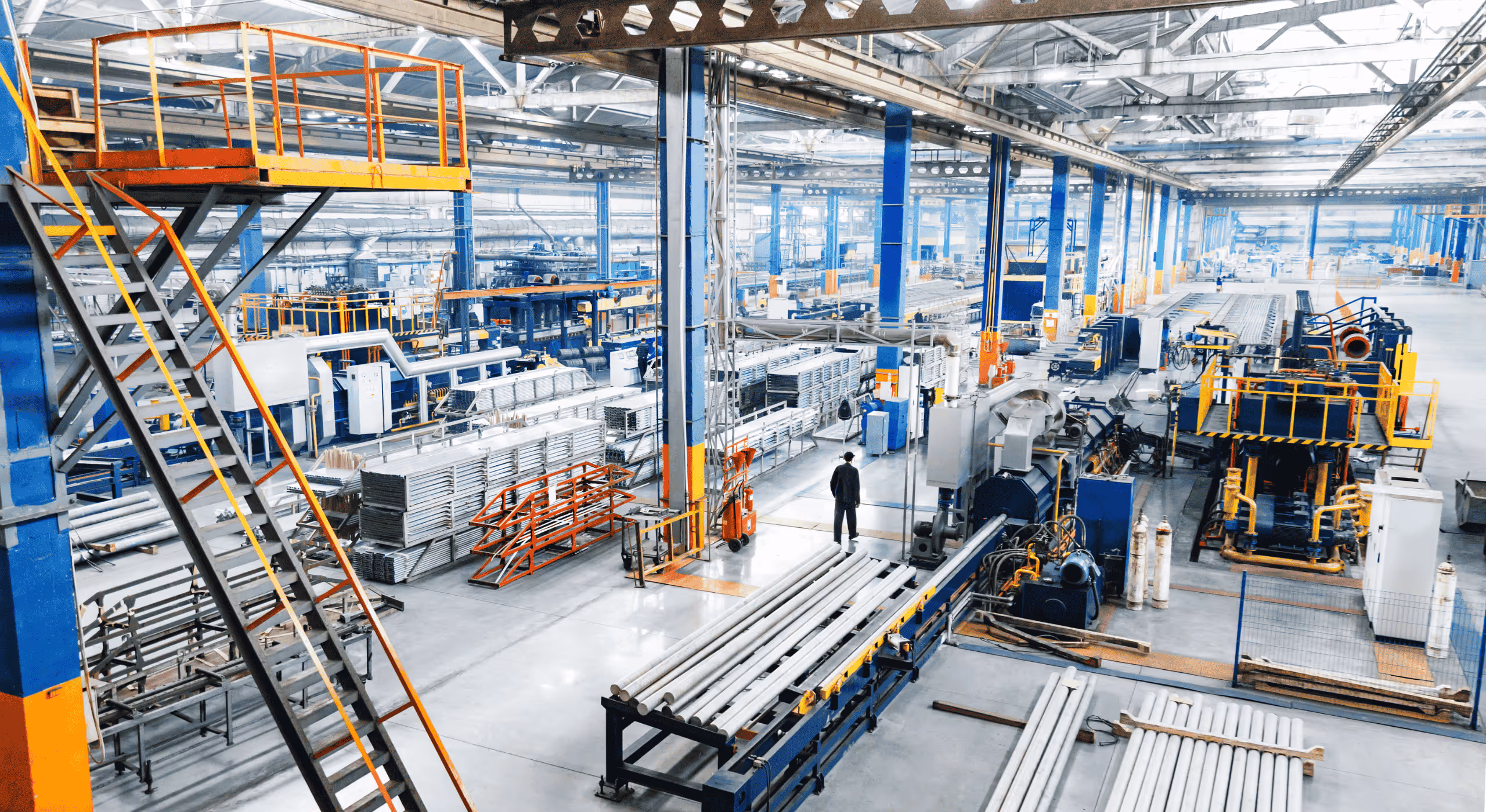 Wide view of a large industrial factory interior with metal pipes, machinery, and a worker in a black uniform.