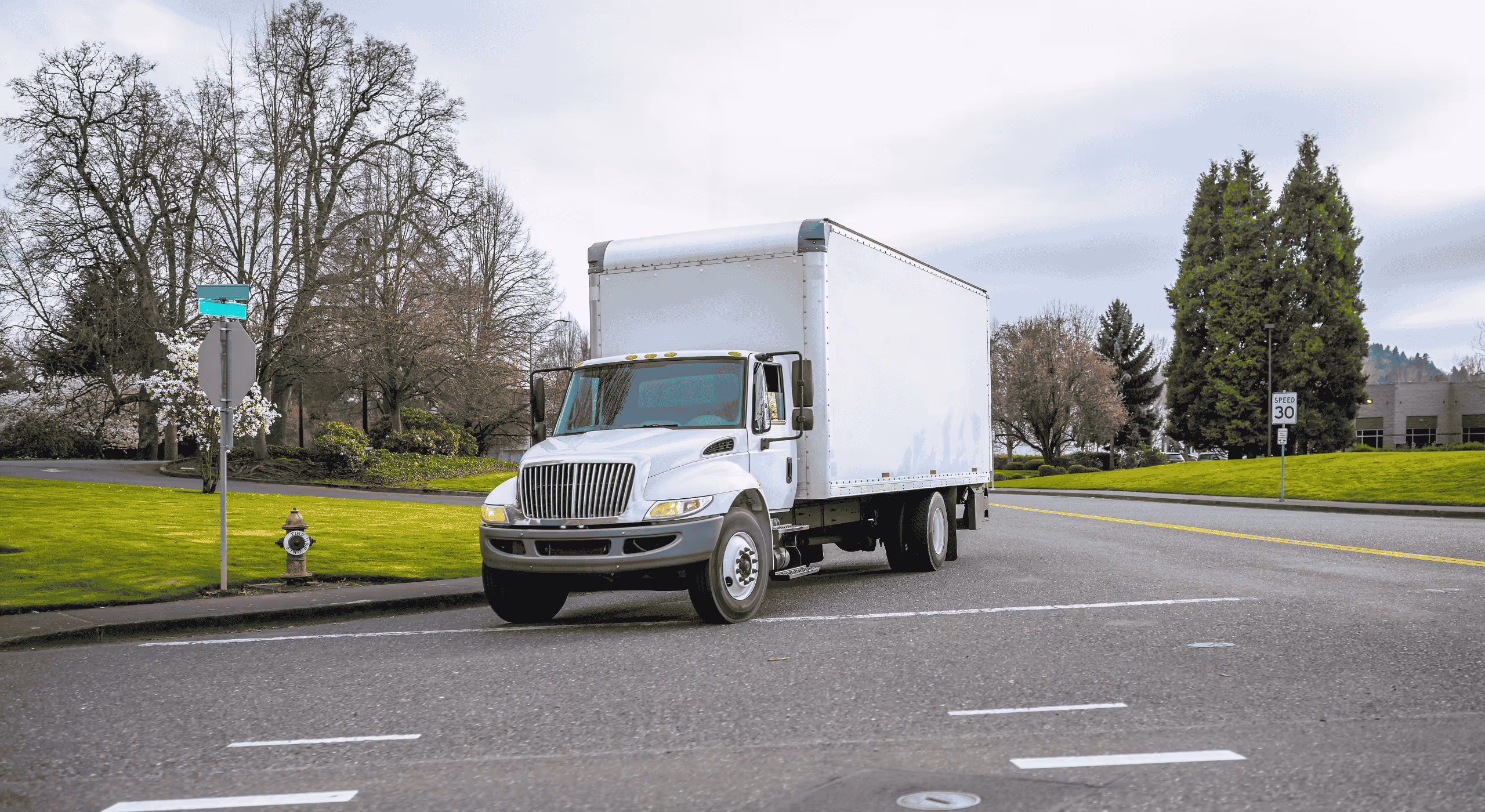White compact semi-truck with a cube box trailer driving on a suburban street near green grass and trees.