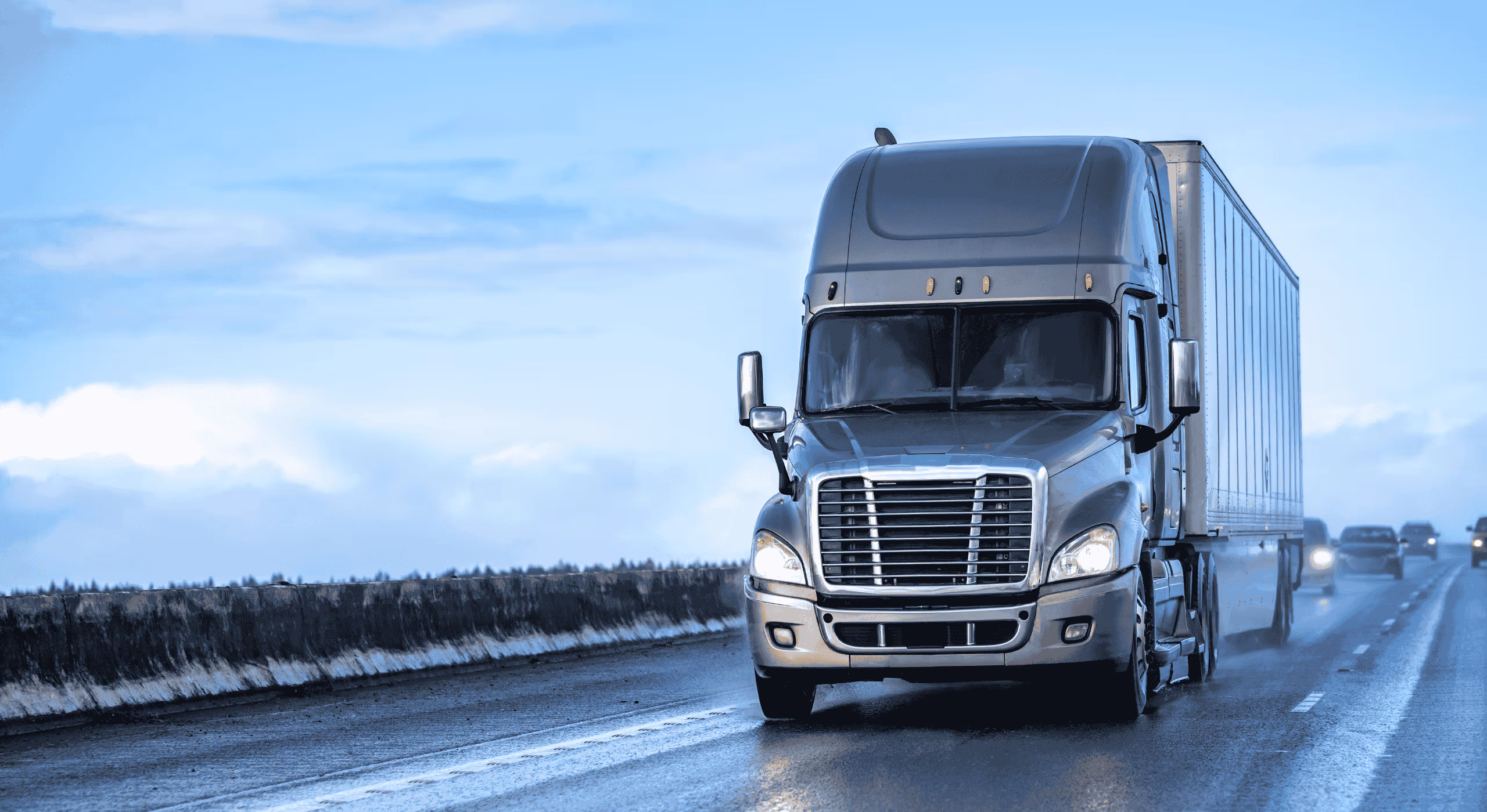 Gray semi-truck driving on a wet highway under a cloudy sky with other vehicles in the background.