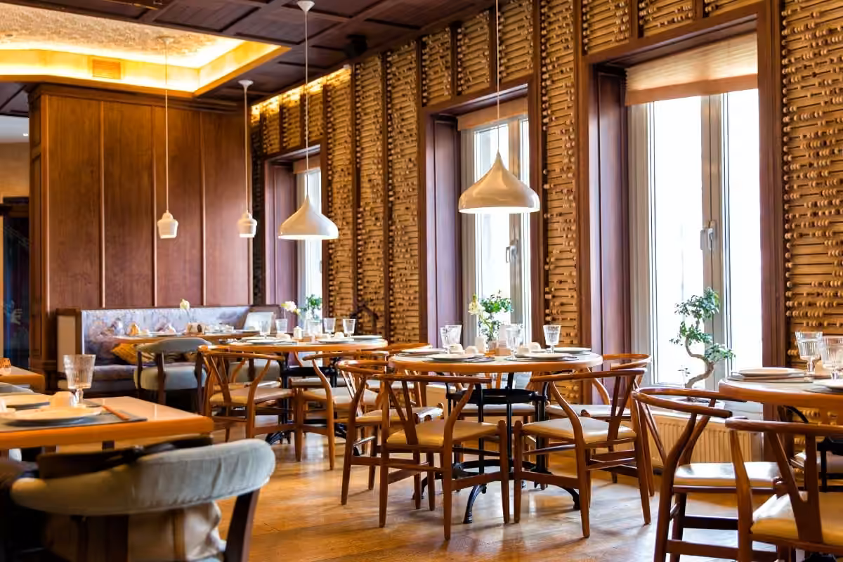 Modern restaurant interior with wooden chairs and tables set with glassware and plates, pendant lights, and large windows letting in natural light.