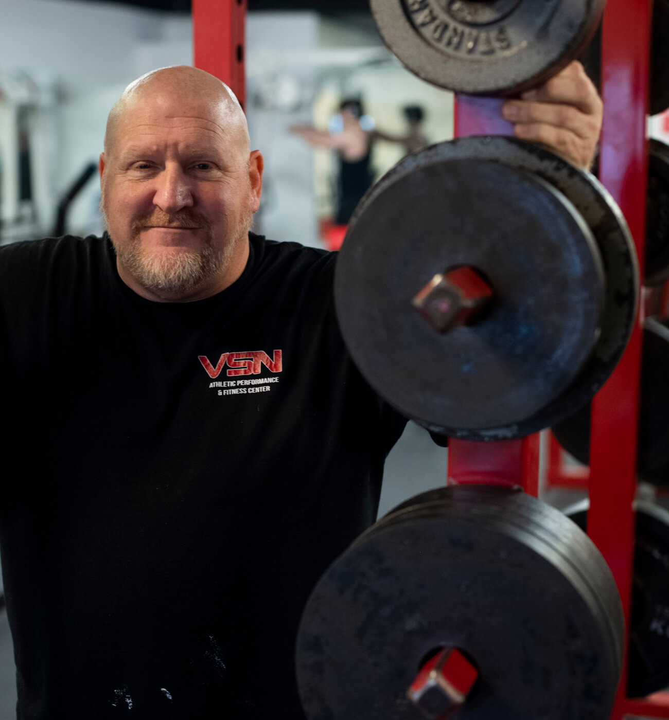 A man smiling in front of a squatting rack. 
