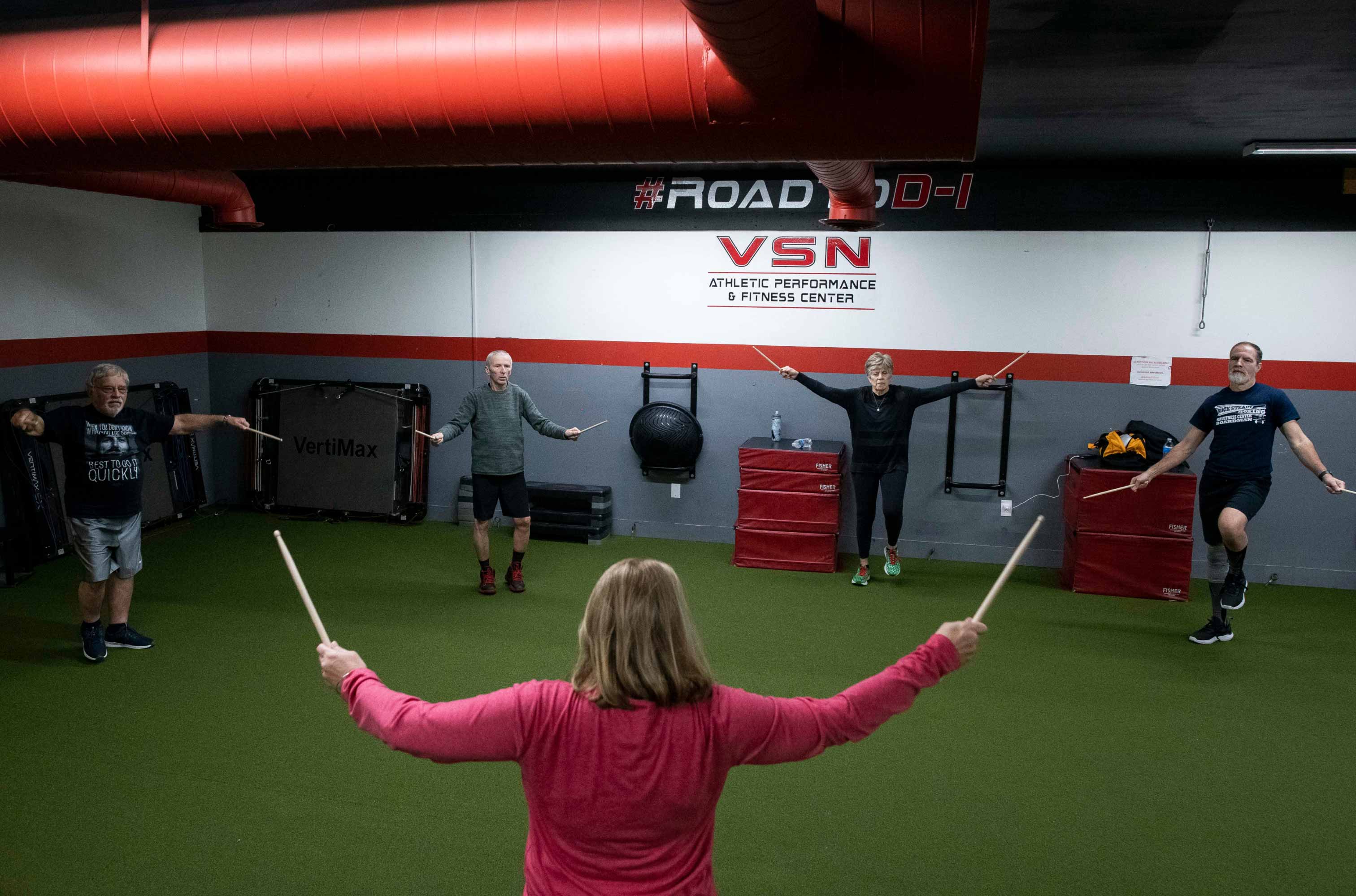 Woman facing boxing class holding up drum sticks. 