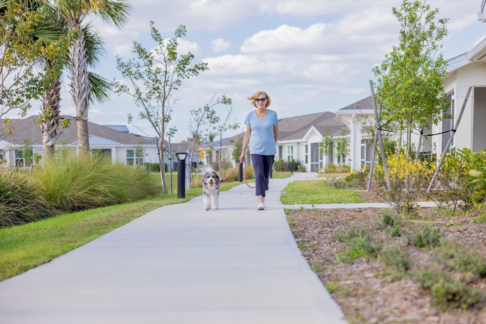 Older adult woman enjoying a walk with her dog along a tree-lined path at Encore at Tradition, surrounded by apartments and villas.