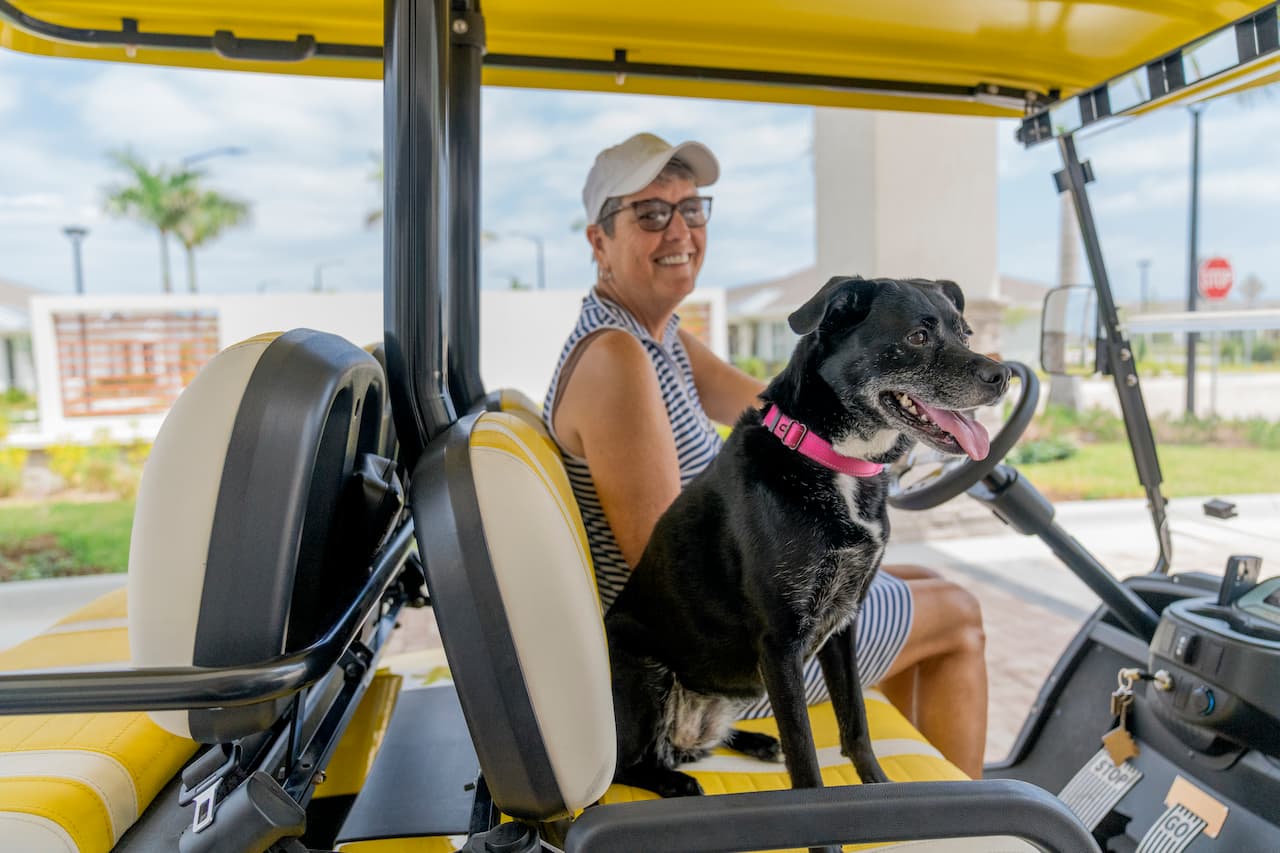 A resident driving her dog in a community golf cart at Encore at Tradition, a pet-friendly apartment in Port St. Lucie, FL, enjoying a sunny day in a 55+ active adult neighborhood.