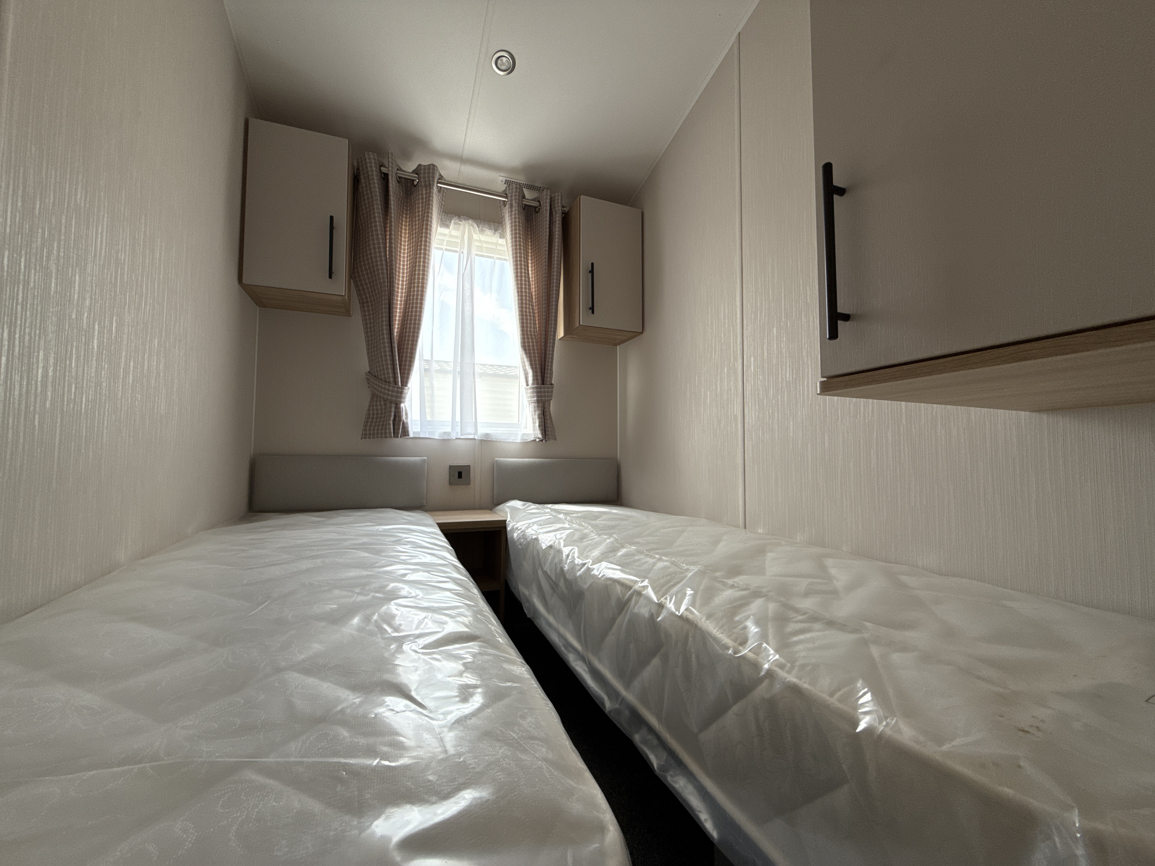Narrow bedroom with two twin beds, curtained window, and overhead cabinets