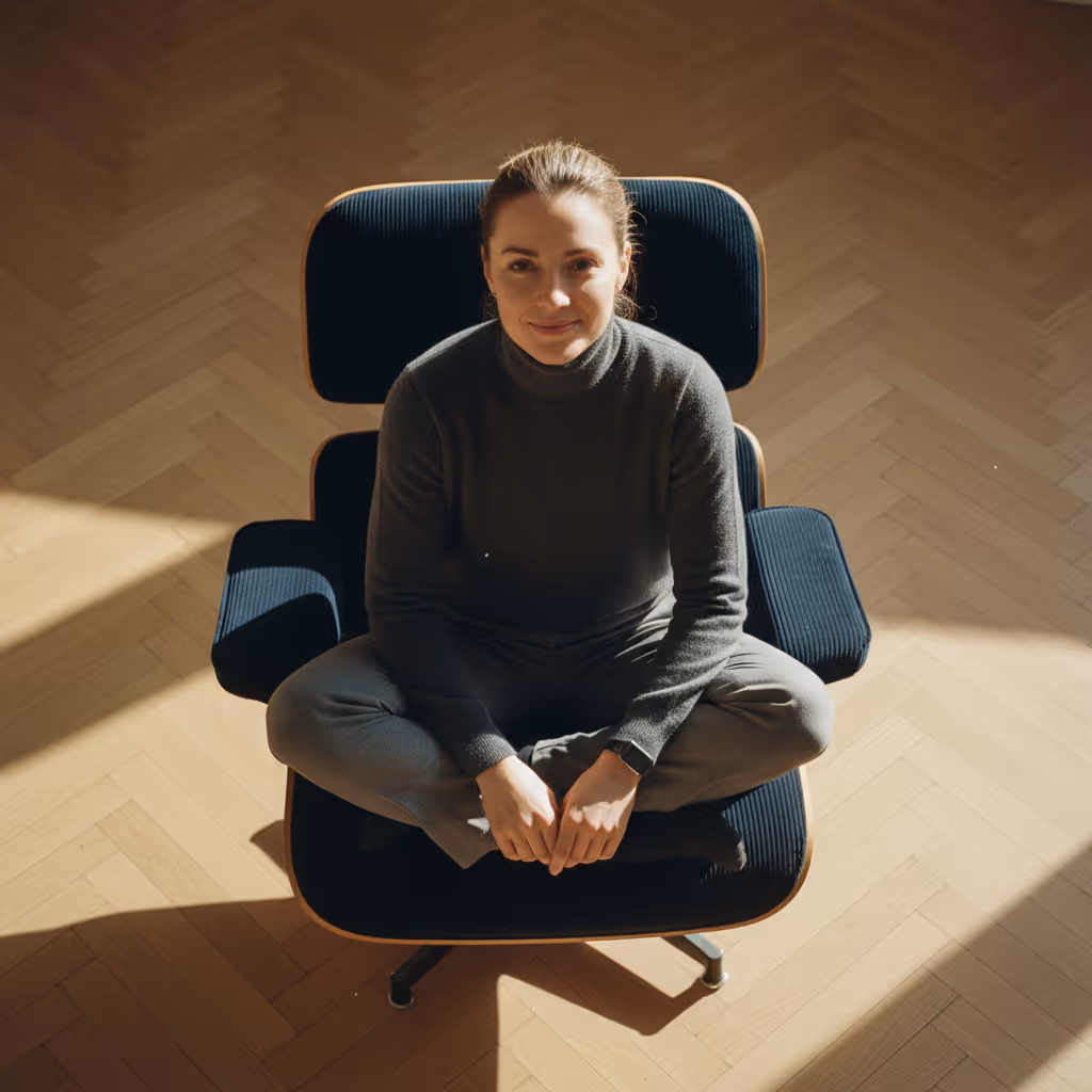 Woman sitting cross-legged on a dark cushioned chair, looking up with a slight smile in a room with wooden flooring.