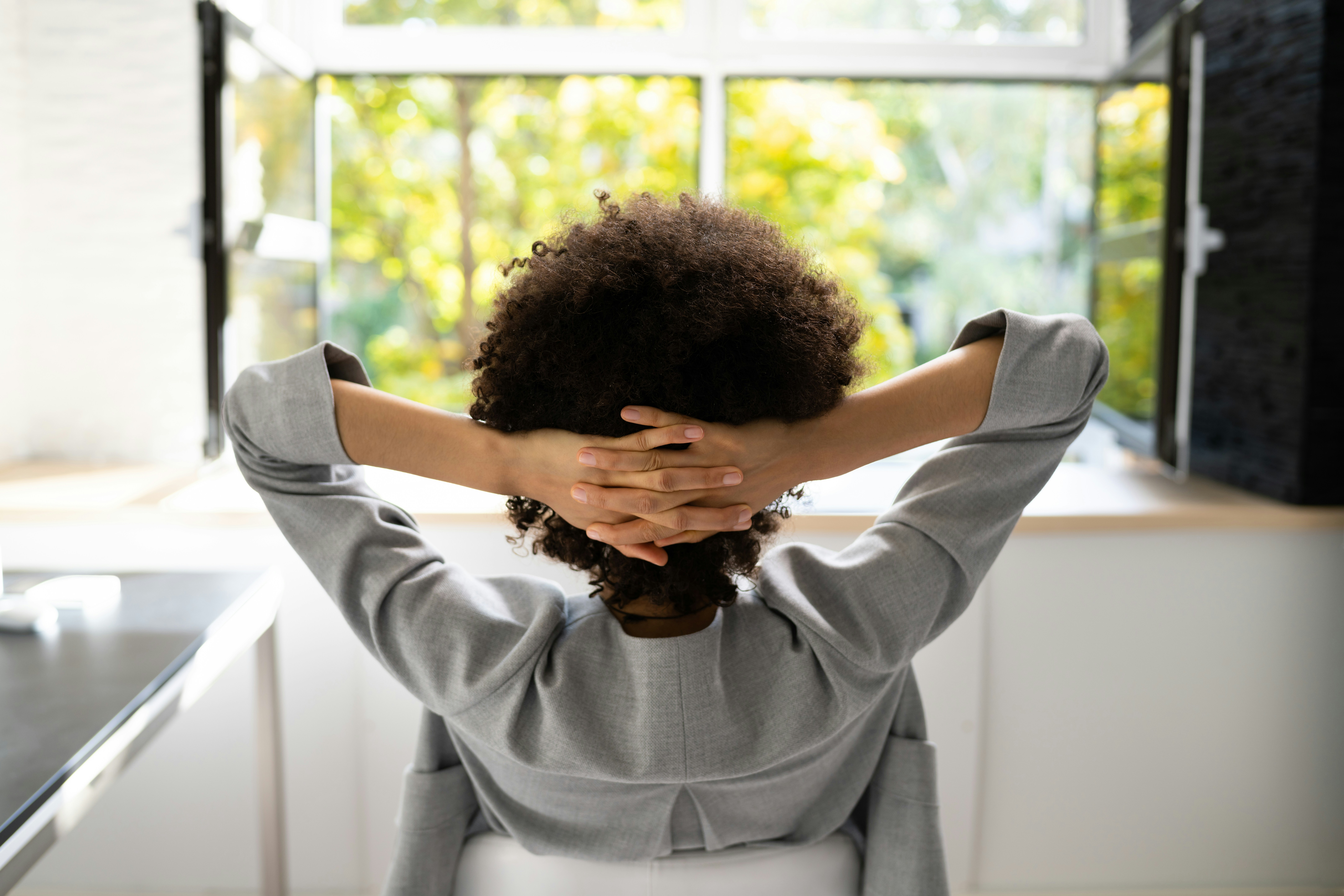 A person leaning back in a chair with hands resting behind their head, gazing out a window into greenery, caught in a quiet pause between moments