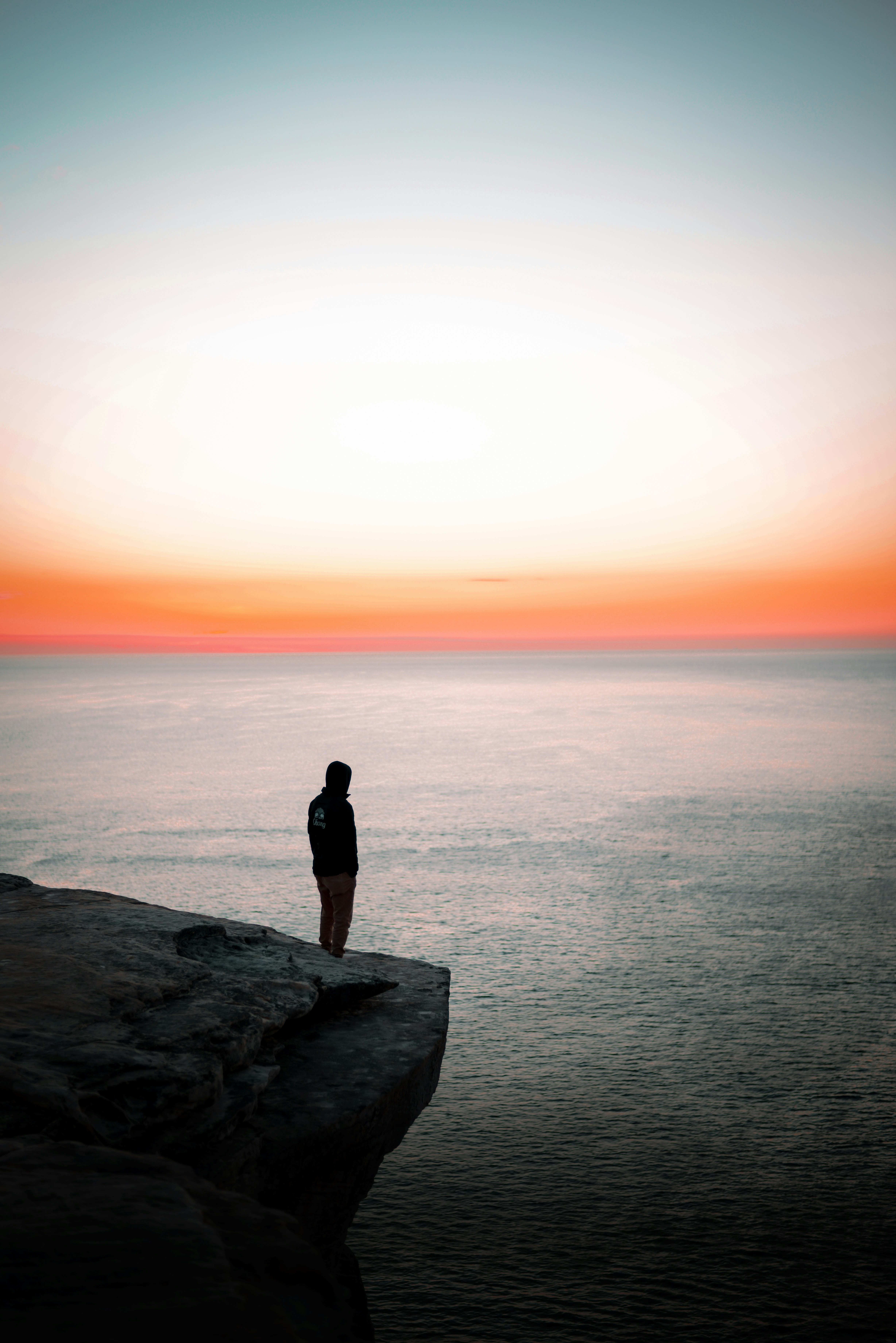 A person standing on a rock cliff at sunset overlooking a calm ocean, the gradient sky shifting from warm orange to deep blue at the horizon