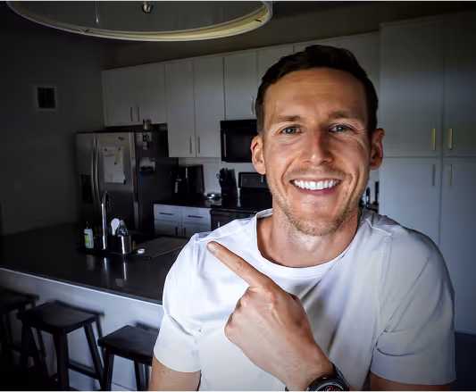 Corey in a white shirt points to himself while standing in a modern kitchen.