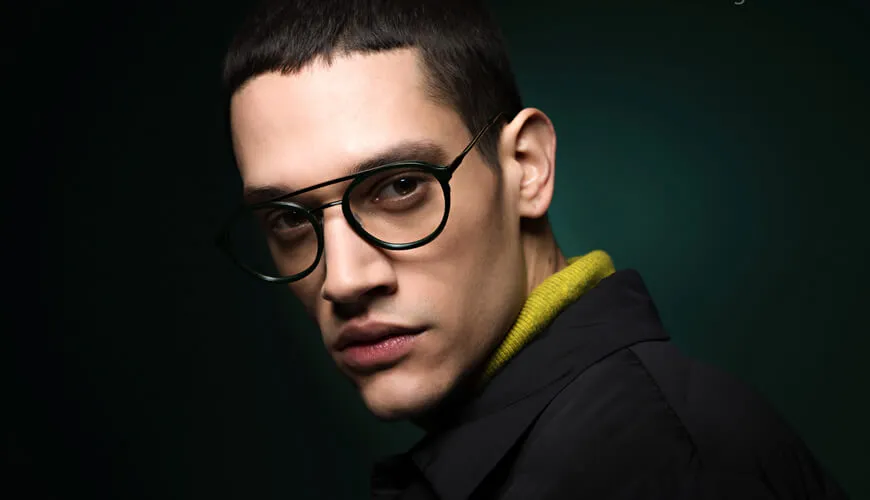 Person with glasses in black jacket and yellow collar against dark background