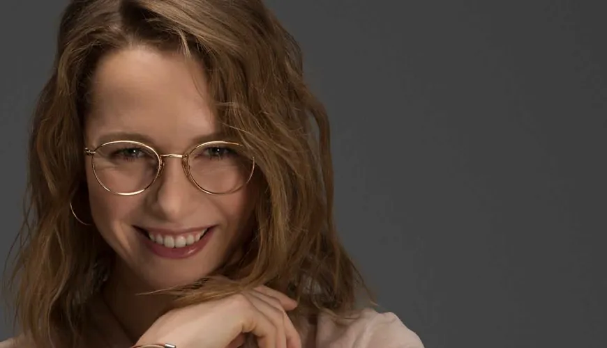 Smiling person with wavy hair and gold-rimmed glasses looking sideways