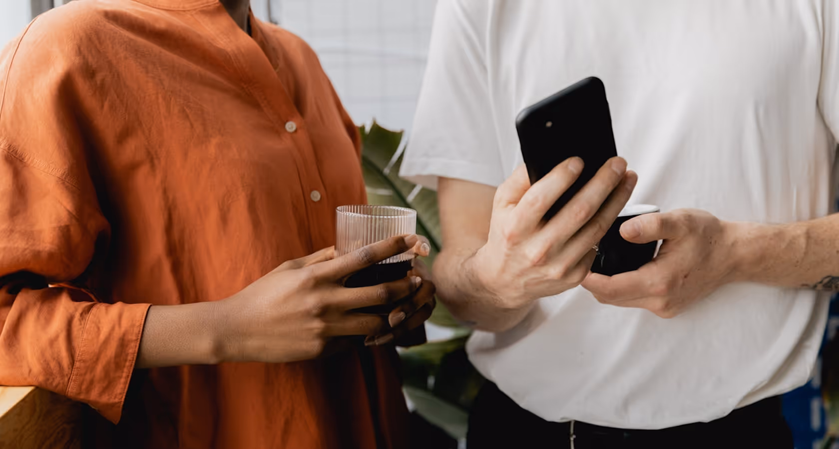 Close-up of two people holding drinks, one person in an orange shirt and the other wearing a white t-shirt while looking at a phone.