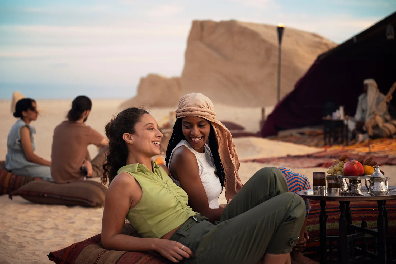Two women smiling and relaxing on cushions in a desert setting with a table of fruit and traditional tea pots nearby.