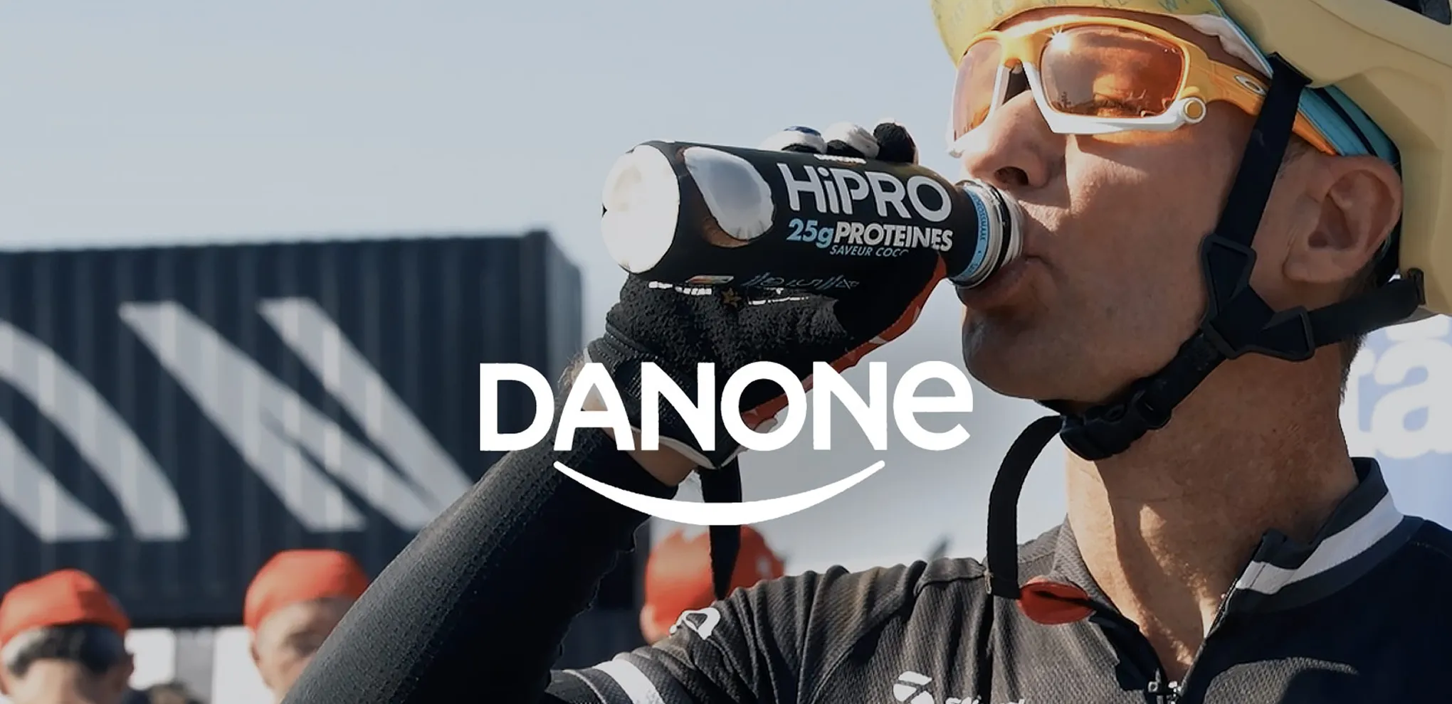 Cyclist wearing helmet and sunglasses drinks from a HiPro 25g protein drink bottle outdoors.