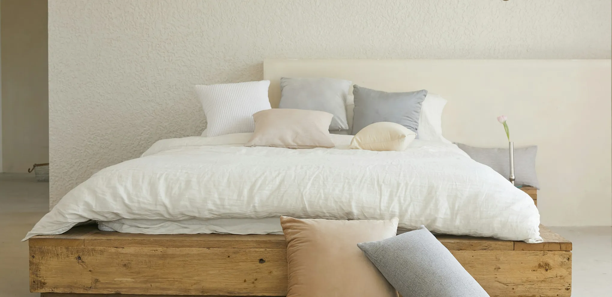 Minimalist bed with white bedding, various neutral-colored pillows, and a single pink flower in a vase on a wooden nightstand.