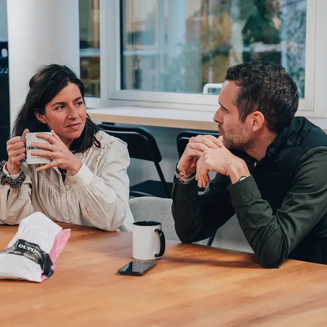A woman and a man sit at a wooden table having a conversation, each with a mug in front of them.