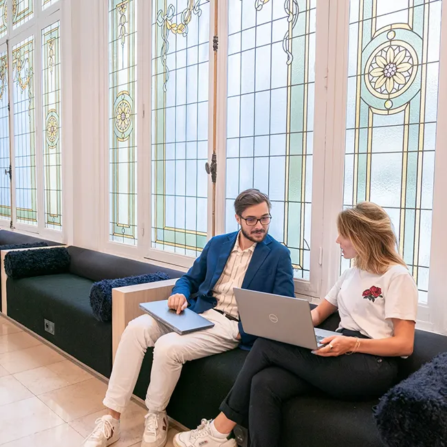 Two people sitting on a black bench in front of large decorative stained glass windows, working on laptops.
