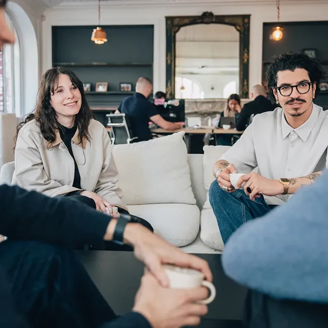 A group of young adults sitting on a couch and chairs in a relaxed office setting, holding coffee cups and smiling during a casual conversation.