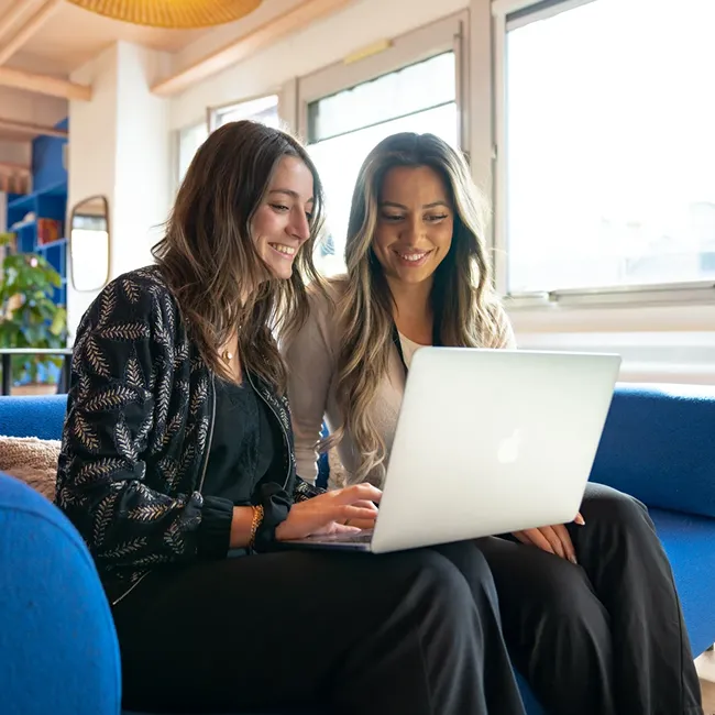 Two women sitting on a blue couch, smiling and working together on a laptop.