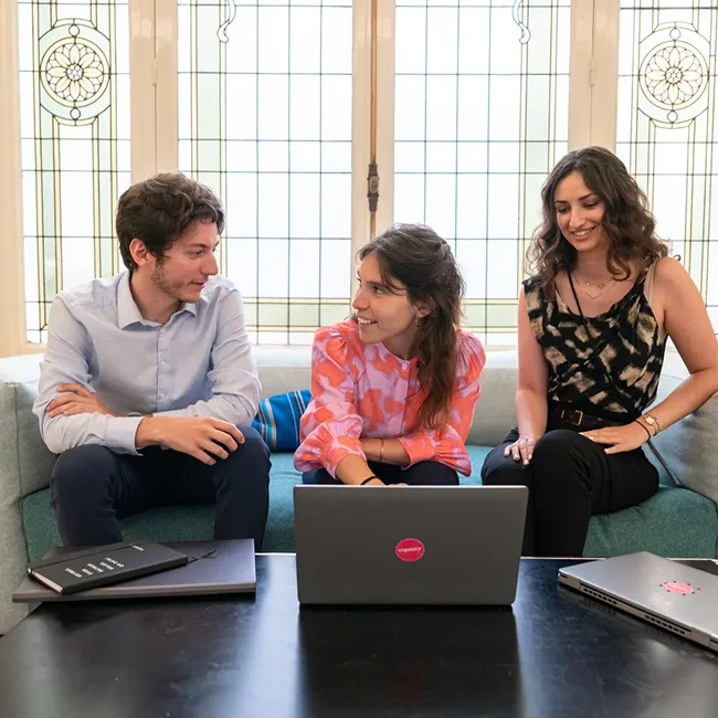 Three young adults sitting on a couch chatting with laptops on a table in front of them.