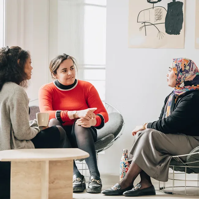 Three women sitting in a bright room engaged in conversation, one wearing a red sweater and another a colorful headscarf.