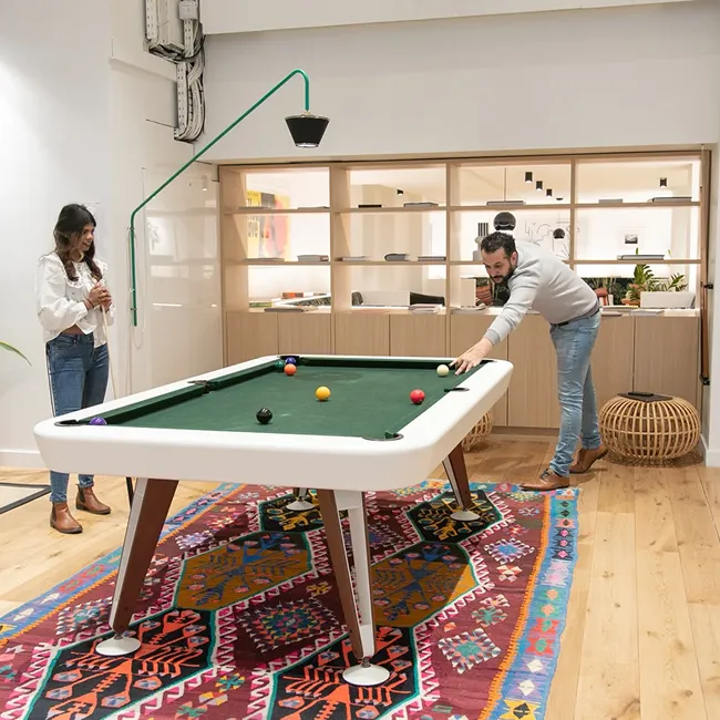 Man aiming to strike a cue ball on a pool table while a woman watches, in a modern room with wooden floor and colorful rug.