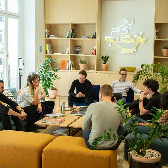 A group of seven people having a casual meeting and conversation in a bright, modern room with plants and wooden shelves, with a neon lamp shaped like a genie lamp on the wall.