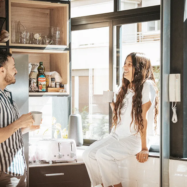 Man and woman enjoying coffee together in a bright kitchen by a large window.