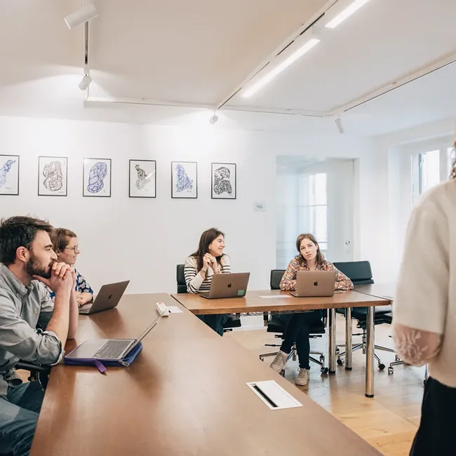 Four people in a bright meeting room listening to a standing person, with laptops open on the wooden tables.