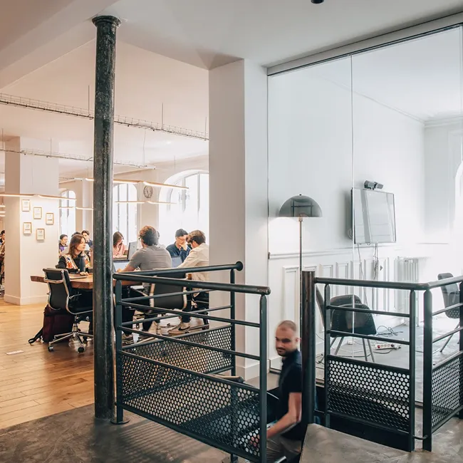 Modern office space with a group of people sitting around a table in a meeting room and a man walking downstairs near a metal railing.