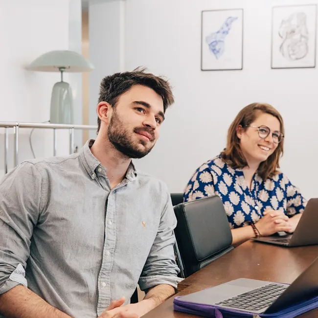 Two young professionals sitting at a table with laptops, one man in a gray shirt and one woman in a blue patterned blouse smiling.