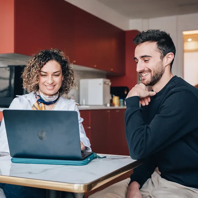 Woman and man sitting at a table looking at a laptop screen, smiling, in a kitchen setting.