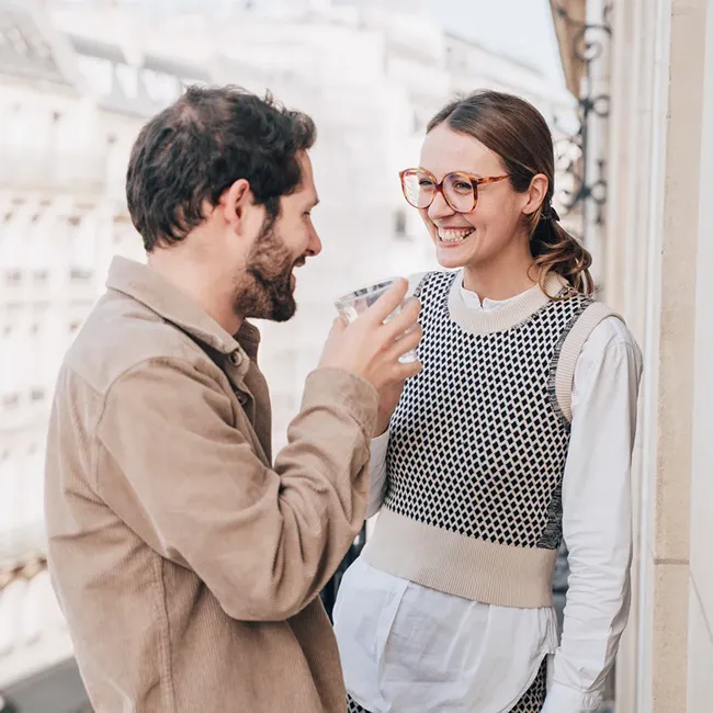 Man and woman smiling and talking on a balcony, the man holding a glass of water.