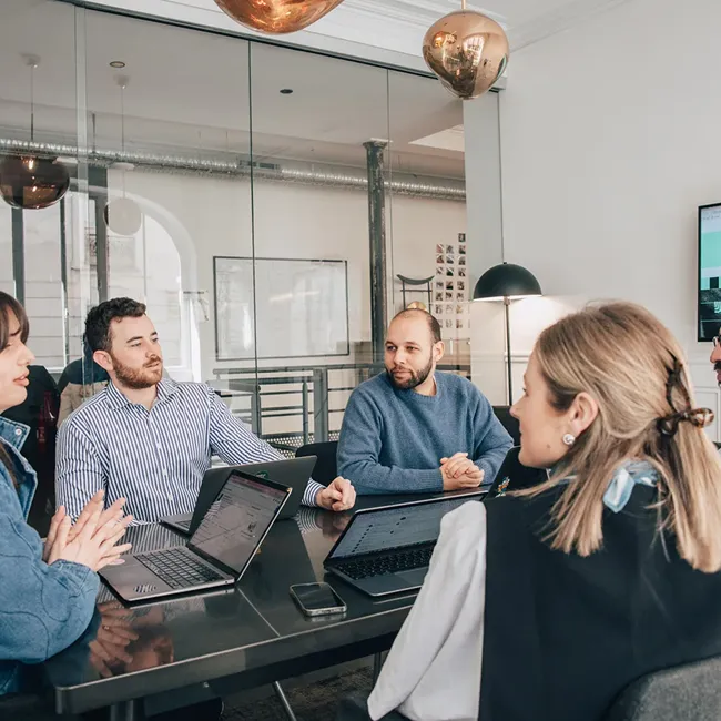 A group of five colleagues having a meeting around a table with laptops in a modern office.