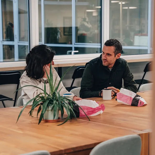 Two people sitting at a wooden table in a modern office, talking over coffee with a potted plant in the foreground.