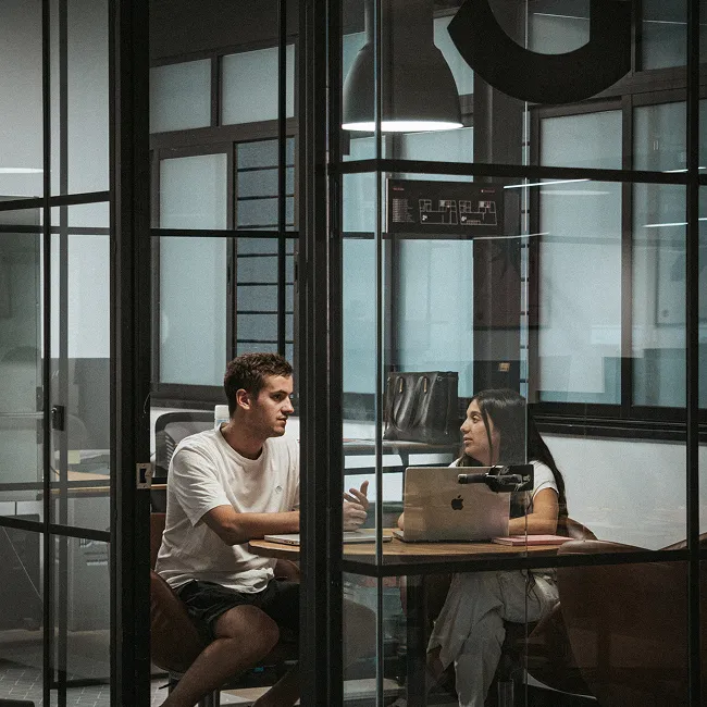 Two people sitting in a glass-walled office room having a discussion with a laptop on the table.
