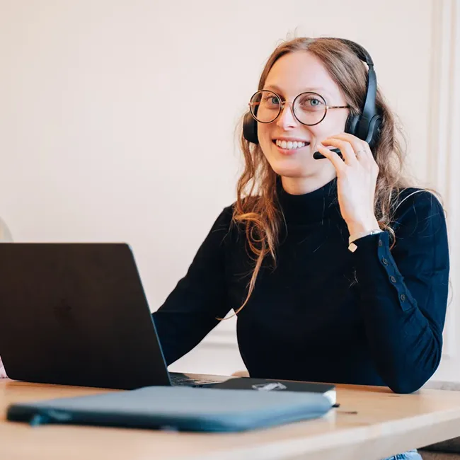 Smiling woman wearing glasses and a headset, sitting at a desk with a laptop and notebook.