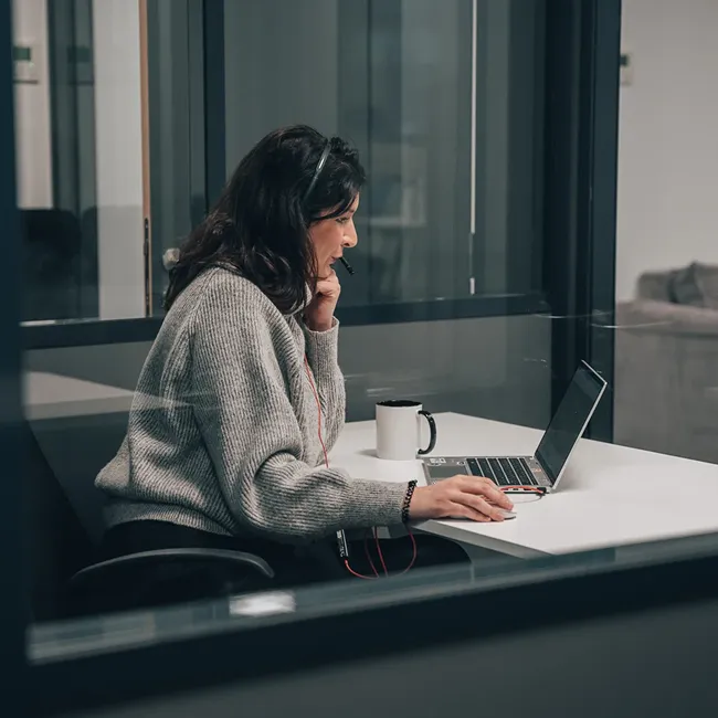 Woman wearing a headset and grey sweater working on a laptop at a white desk with a coffee mug.