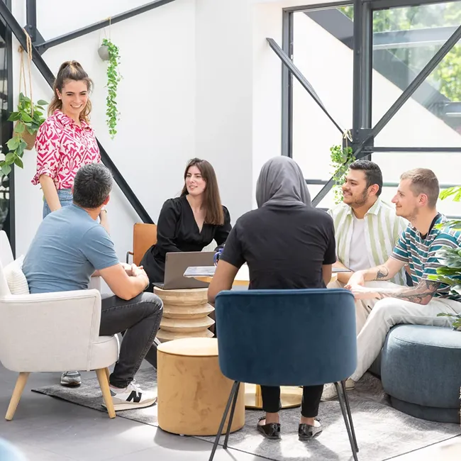 Five people having a casual meeting in a bright, modern office with large windows and plants.