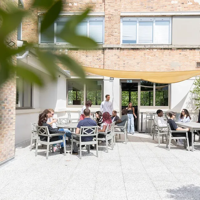 Group of people sitting and standing around outdoor tables in a sunny courtyard with brick walls and large windows.