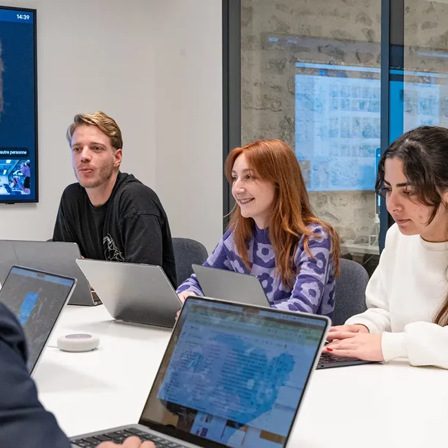 Three people sitting at a conference table with laptops, engaged in a meeting or discussion in a modern office.