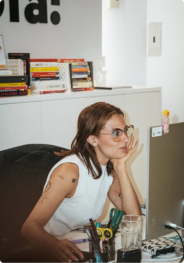 Woman with glasses and tattoos working at a desk with a computer, surrounded by office supplies and books.